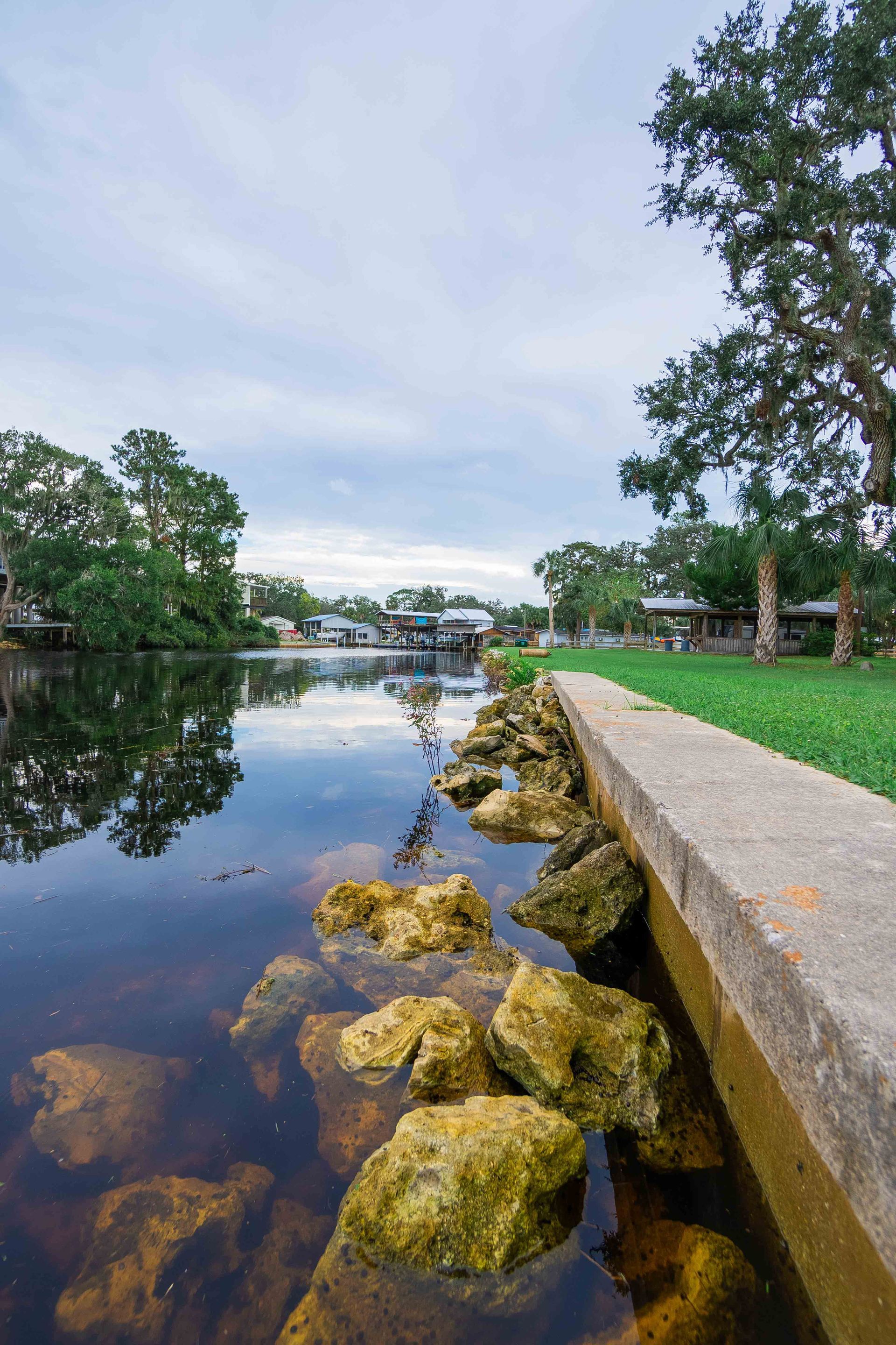 Riverbank with rocks, concrete edge, and trees reflecting on calm water under a cloudy sky