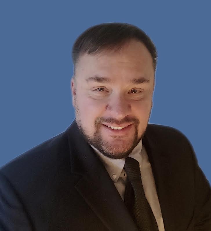 Man in a suit and tie smiles at the camera against a blue backdrop.