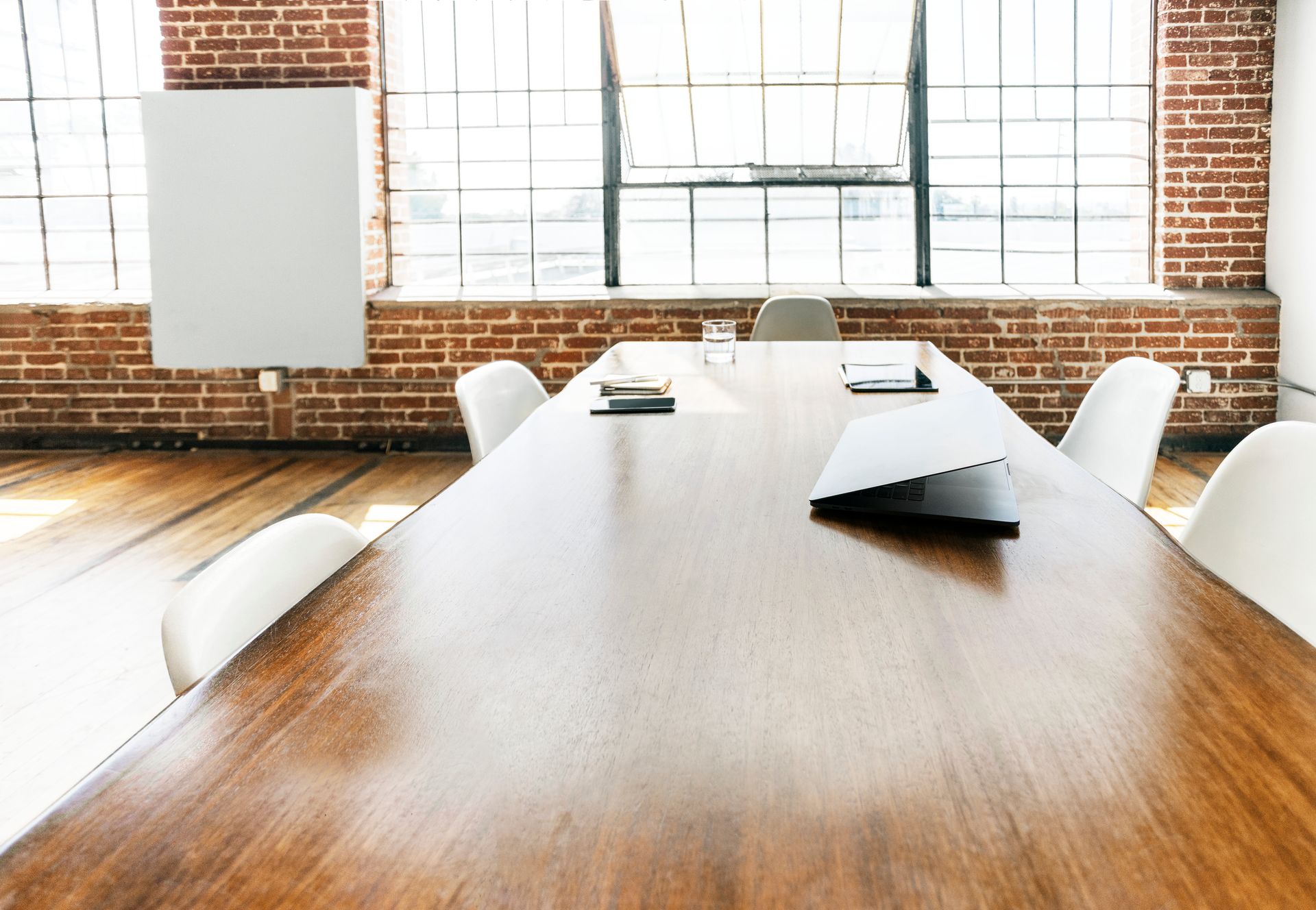 Long wooden conference table in a room with exposed brick walls, a large window, and white chairs.