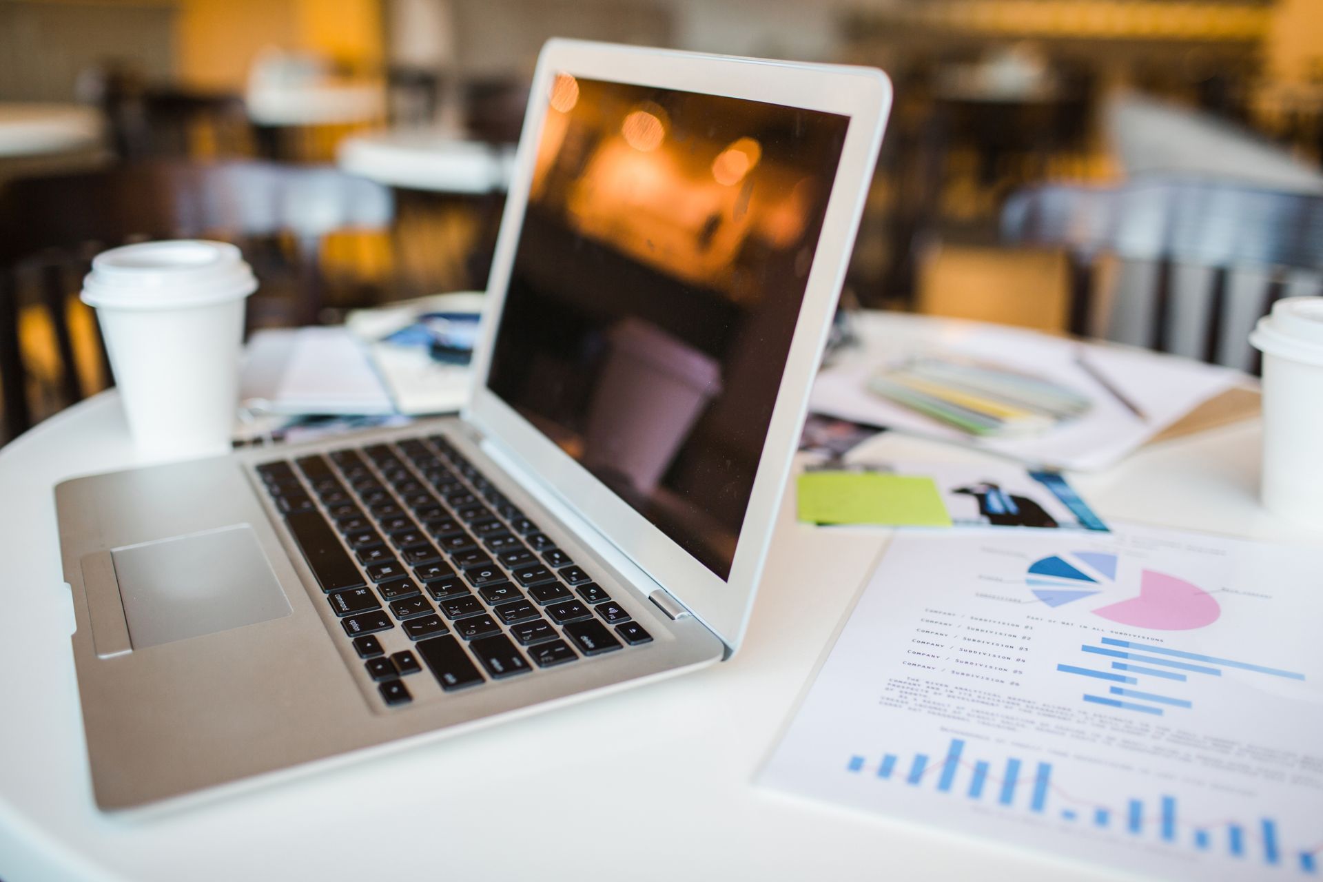 Laptop, coffee cups, and charts on a white table in a cafe setting.