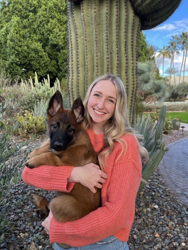 Smiling woman in a coral sweater holding a brown puppy in a cactus garden