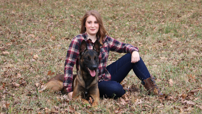 Woman sitting outdoors on grass with a German shepherd, both looking at the camera.