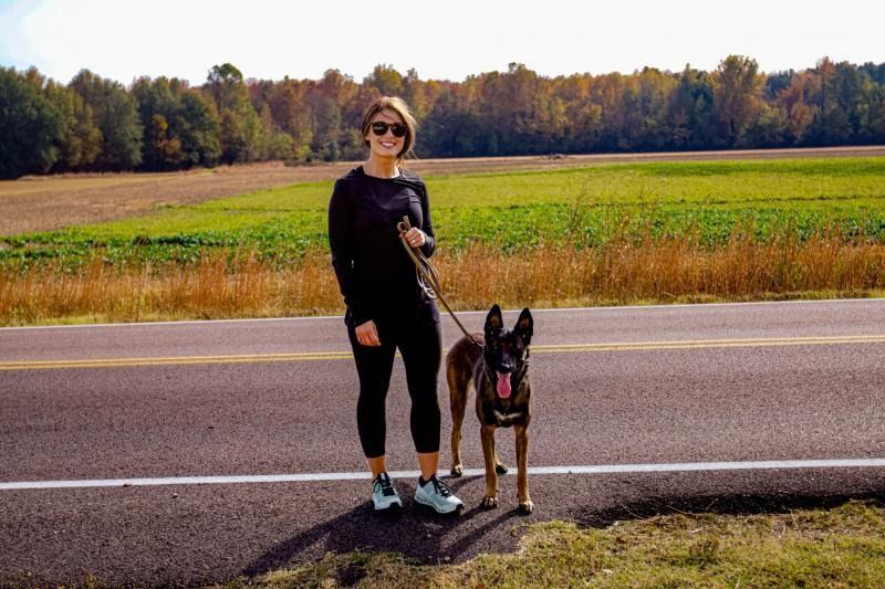 Woman standing with a dog on a roadside beside a grassy field and trees