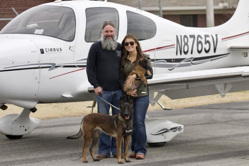 Two people and a dog standing in front of a small white airplane on the tarmac