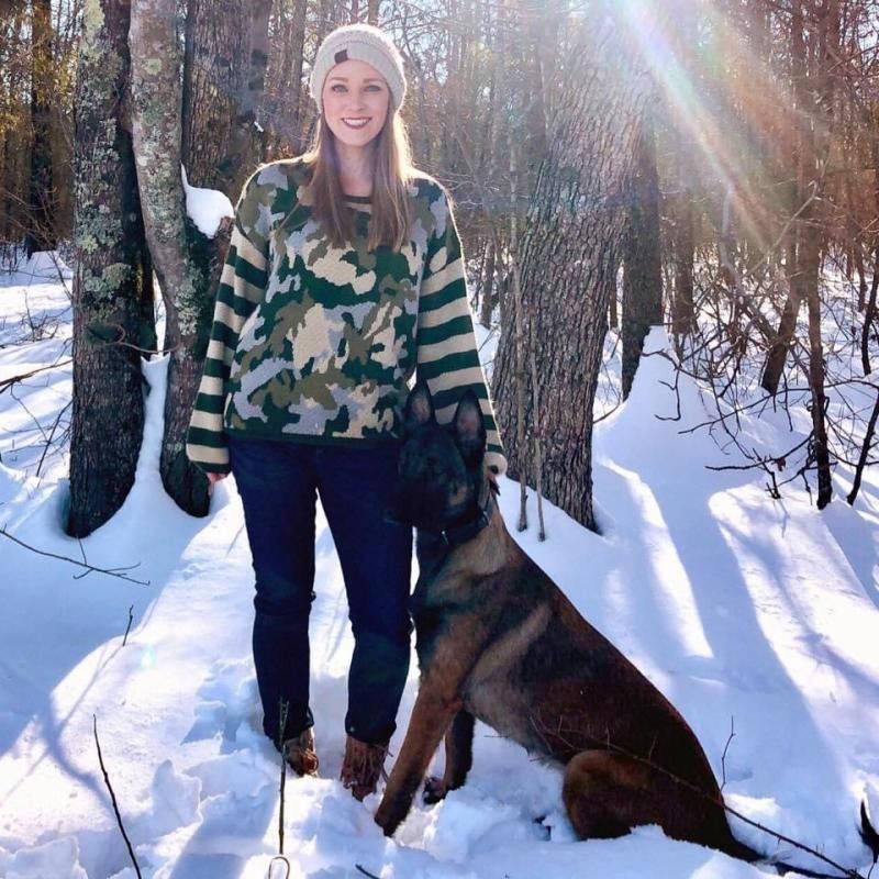 Woman standing in snowy woods with a dog beside a tree in bright sunlight