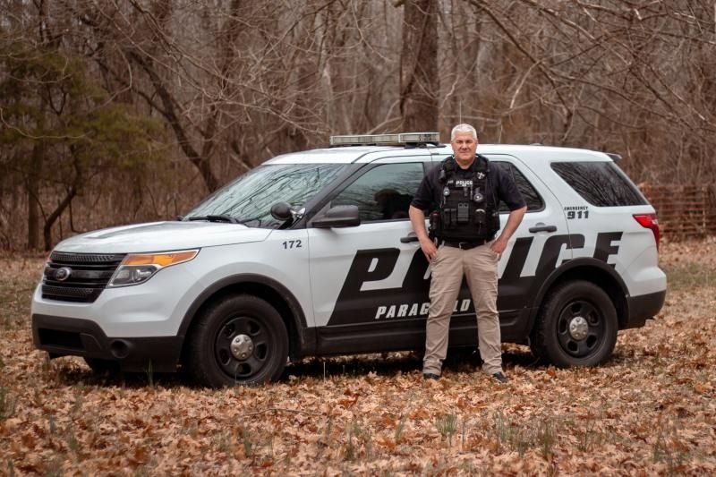 Police officer standing beside a marked SUV in a wooded area in autumn