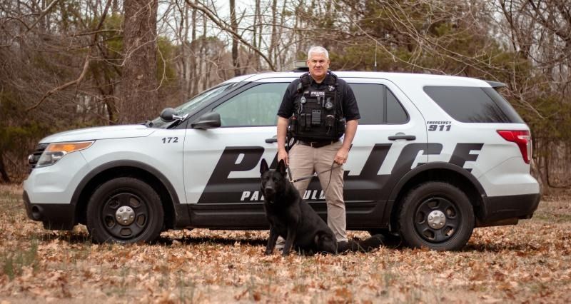Police SUV in a wooded area with an officer standing beside a black police dog.