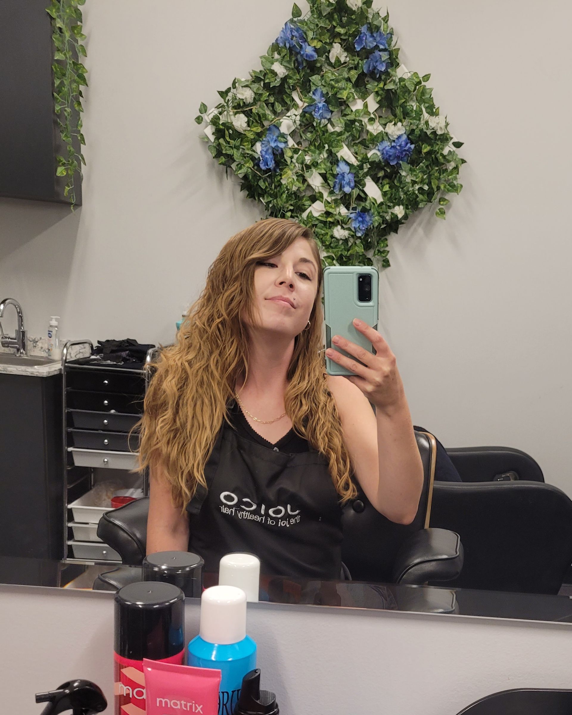 Woman in salon taking selfie, curly hair, holding phone. Decorative floral arrangement on the wall.