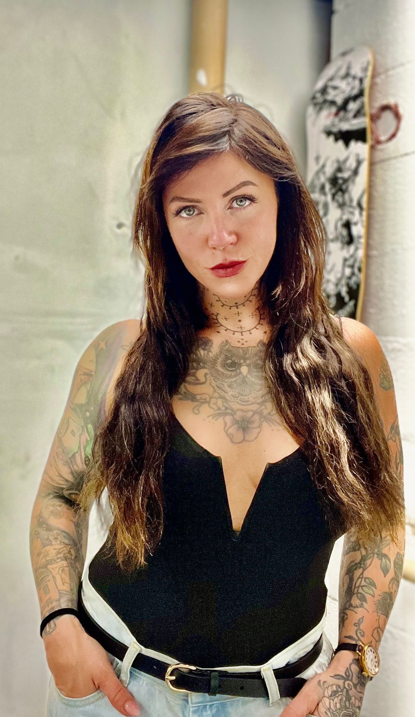 Woman with long dark hair and tattoos posing indoors, wearing a black top and light jeans.