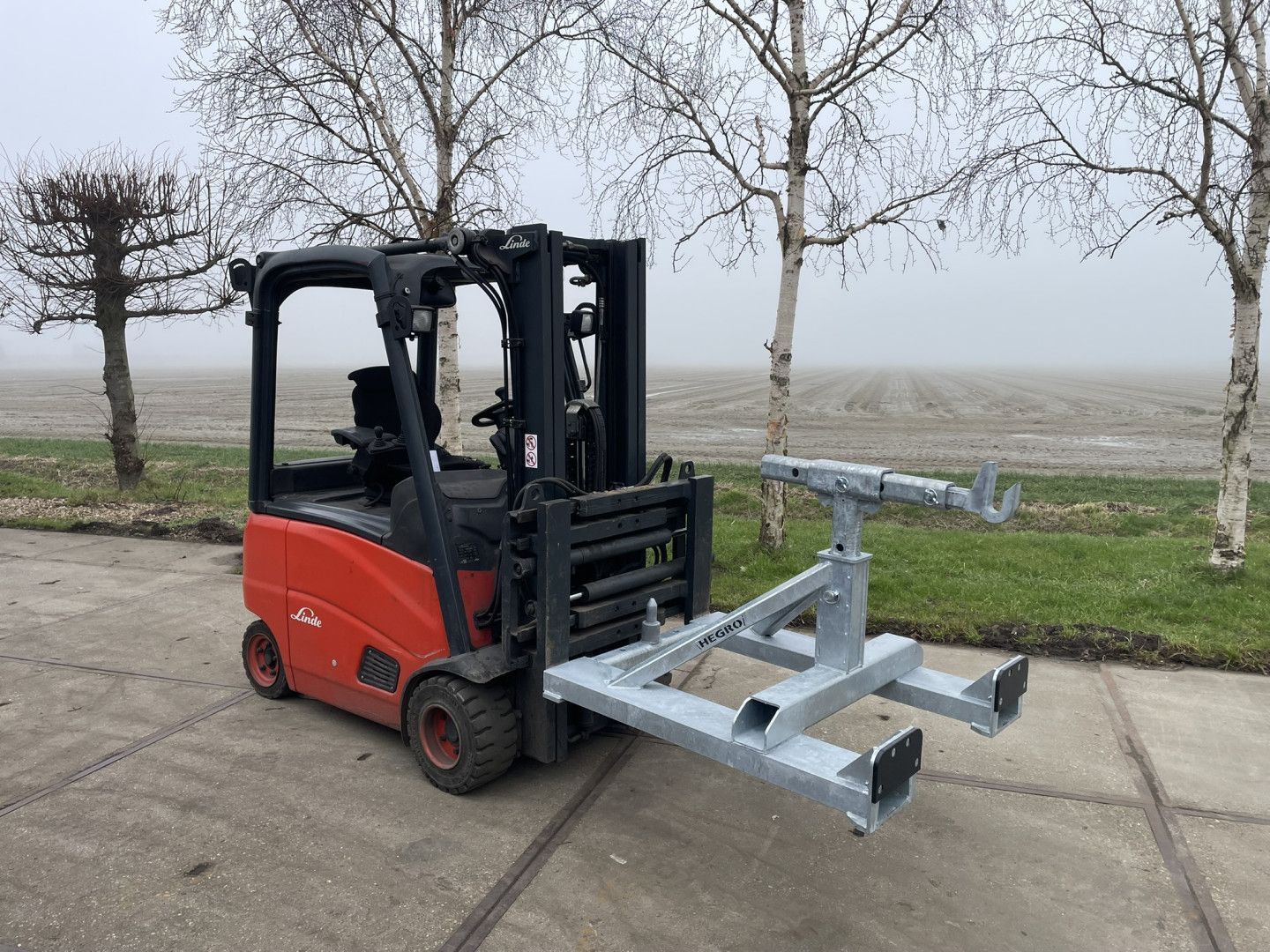 Rode heftruck met een metalen frame-aanbouwdeel op een mistige wegberm met kale bomen.