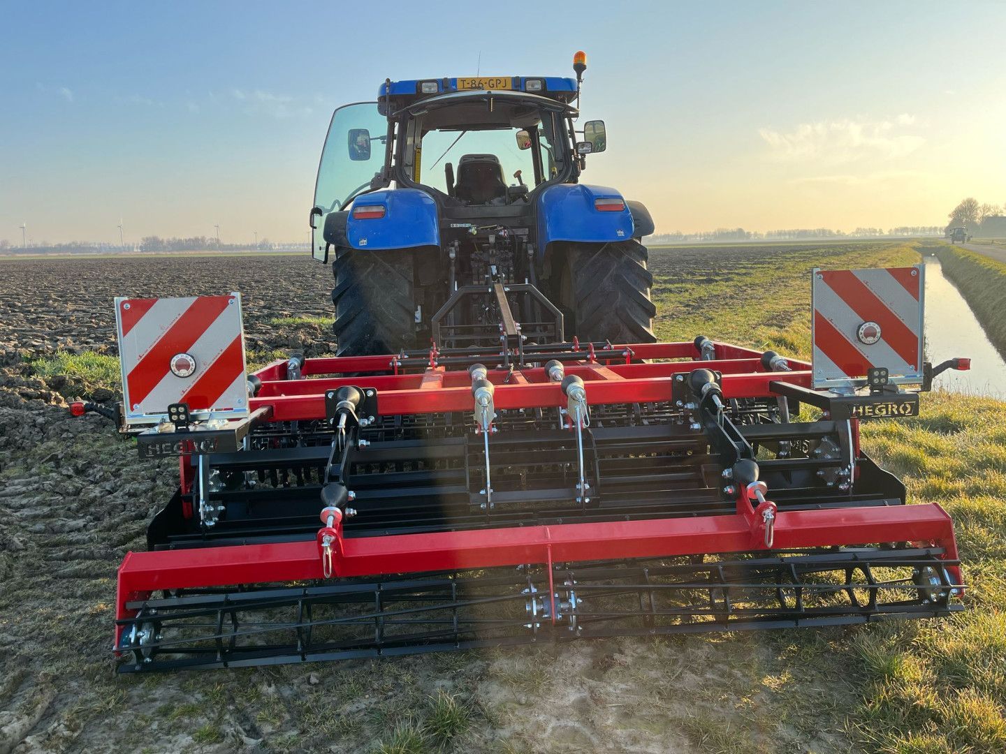 Blauwe tractor met rode zaaibedbereider in een geploegd akkerland bij zonsondergang.