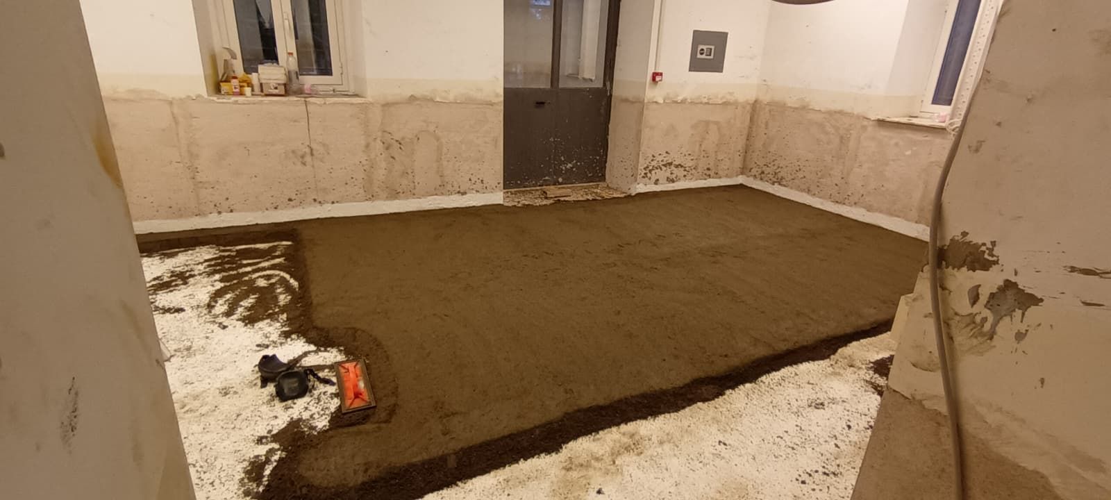 Stanza con pavimento in cemento fresco, pareti grezze e una porta sullo sfondo. Cantiere edile.