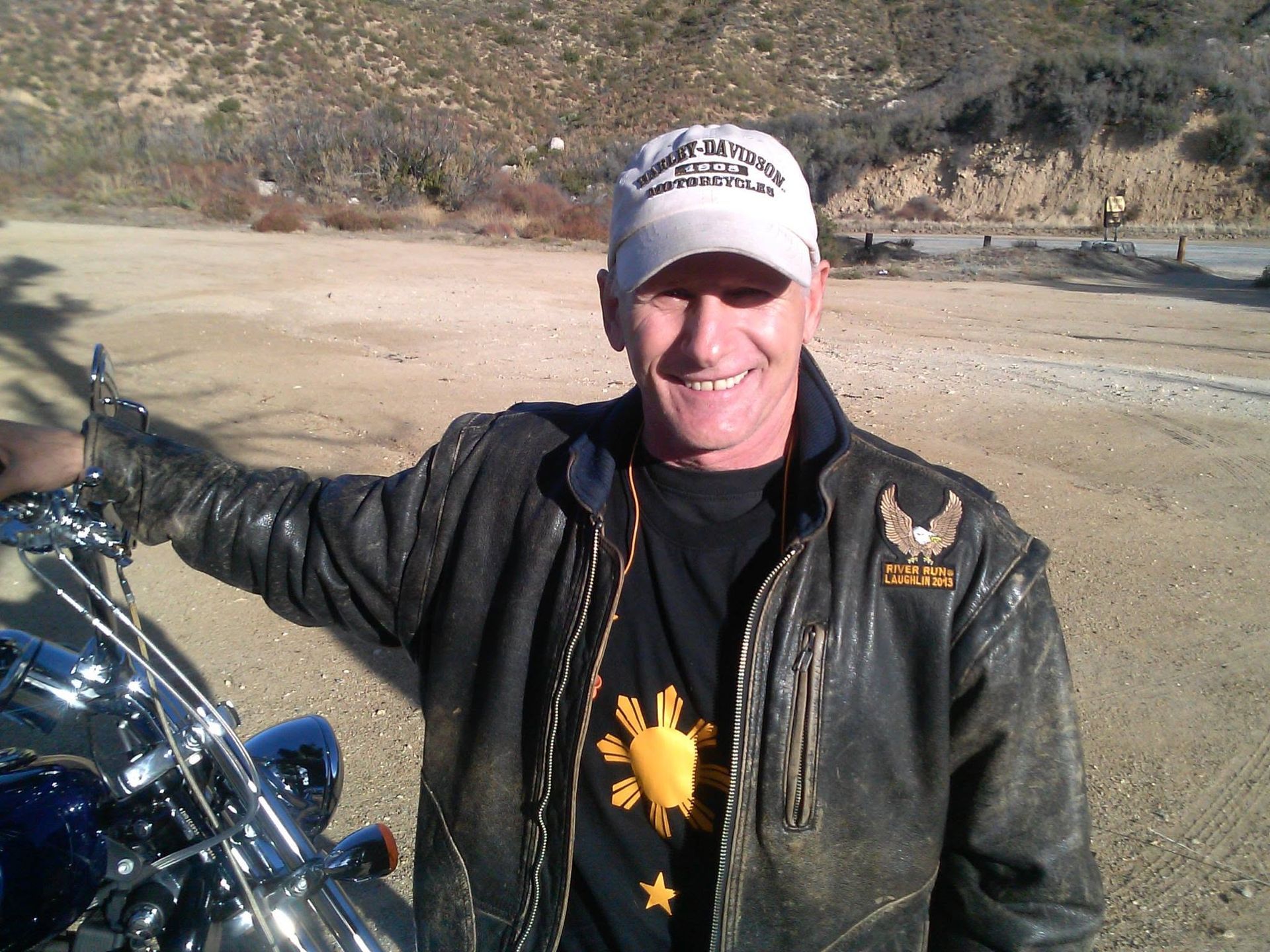 Smiling person in a black jacket and cap standing beside a motorcycle in a desert landscape