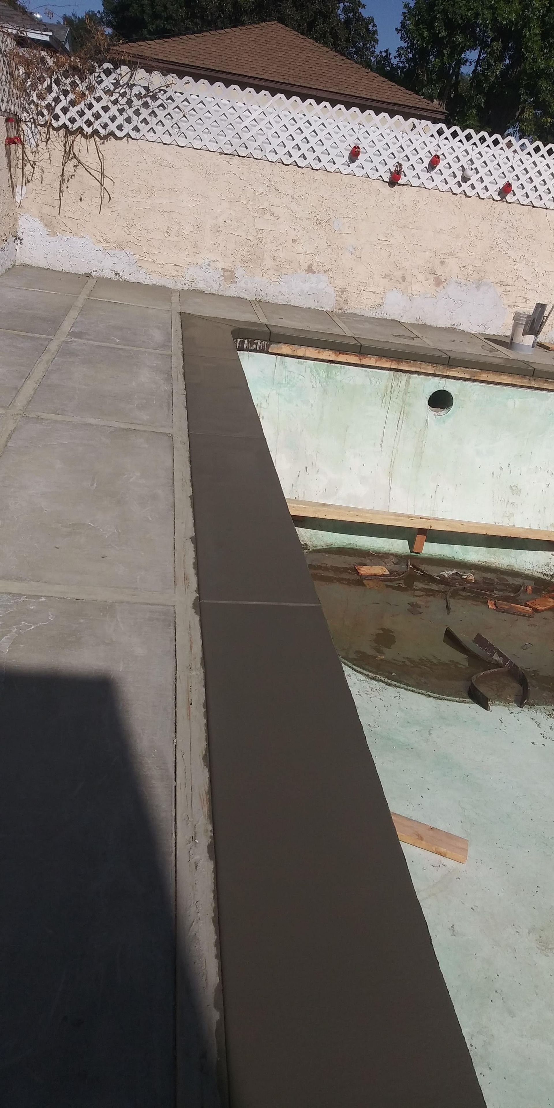 Top-down view of a patio edge beside a tiled roof and glass panel, with a white wall and outdoor debris.