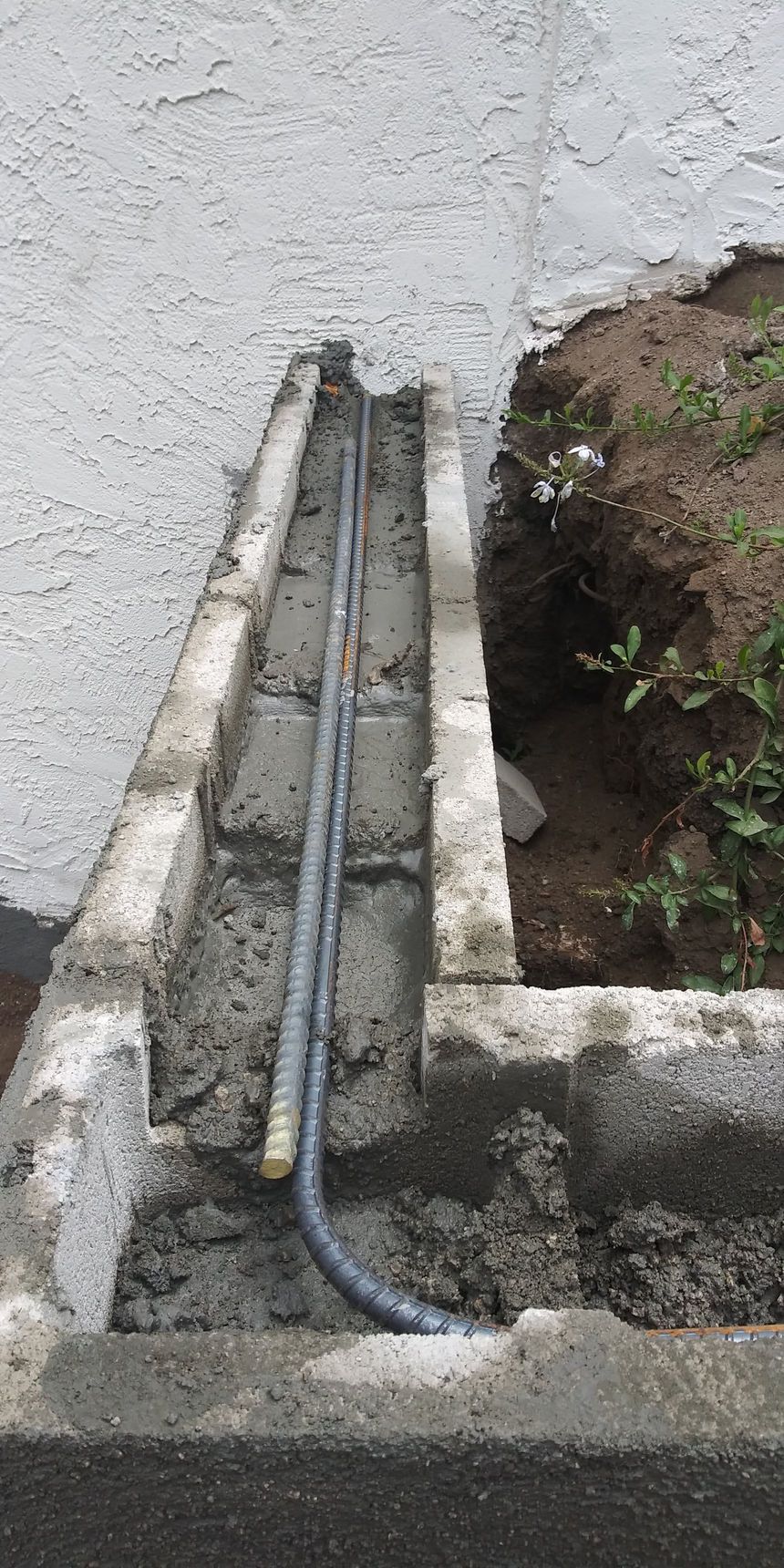 Two metal pipes run through a narrow concrete trench beside a white wall and exposed soil.