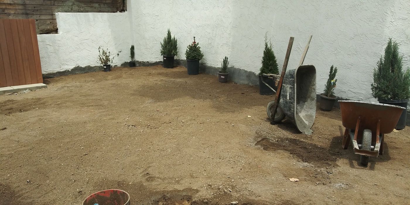Gravel yard beside a white wall with small potted conifers and garden tools near a wheelbarrow