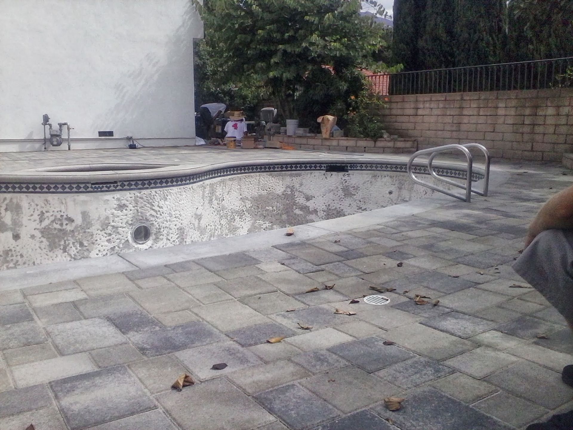 Empty outdoor pool with a tiled deck, metal ladder, and a stone wall in the background.