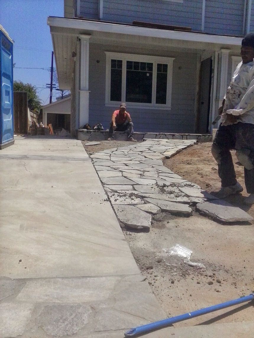Cracked concrete path beside a house, with a worker crouching near the entrance and a man standing at right.