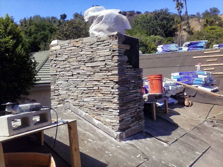 Stacked stone chimney under construction on a rooftop, with tools and materials nearby.