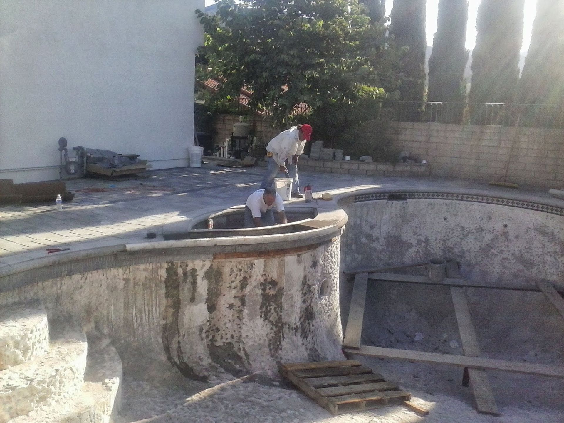 Empty outdoor swimming pool with a person standing on the curved edge near trees and a wall