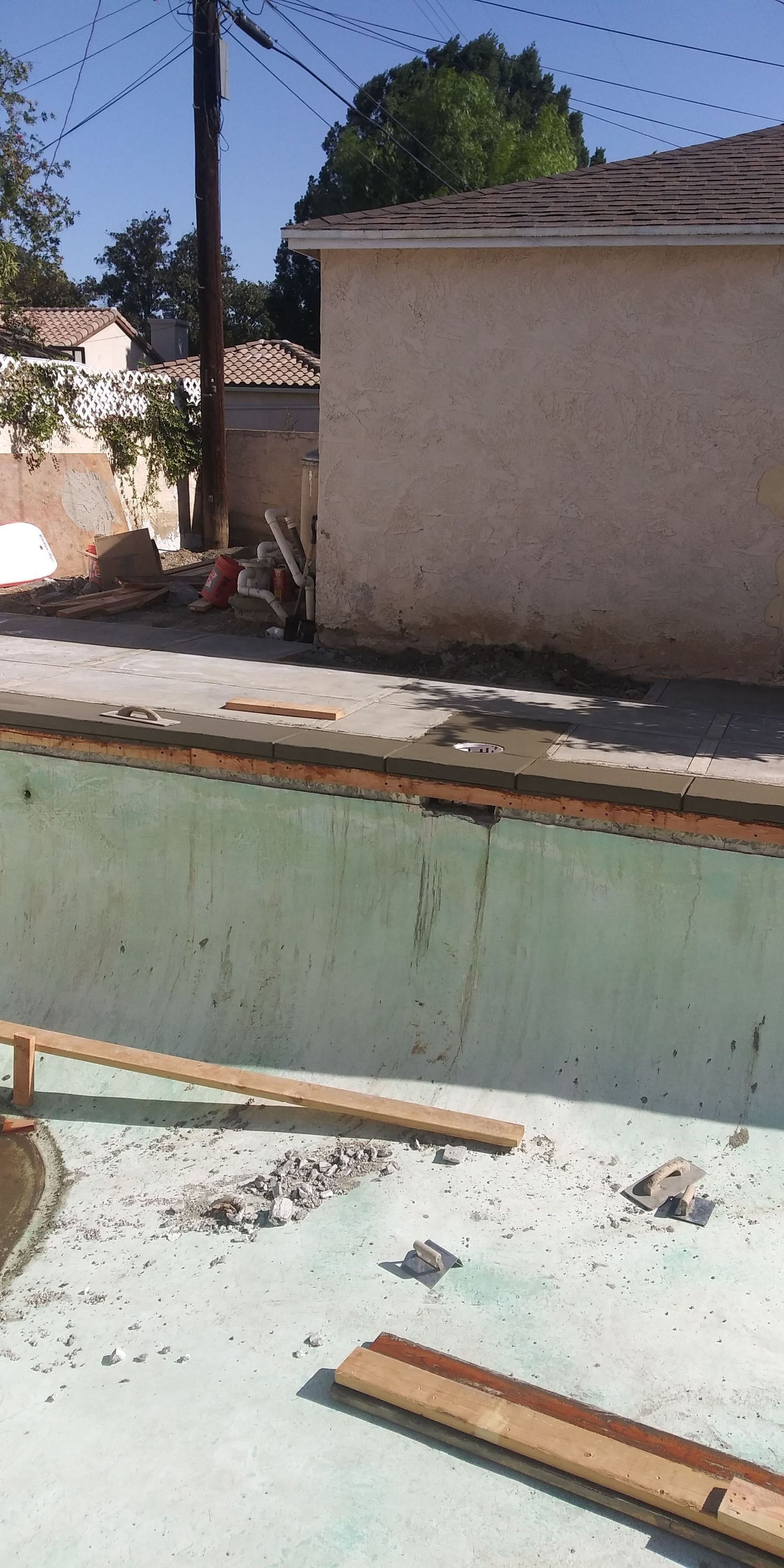 Empty swimming pool with debris, beside a beige wall and trees in bright sunlight