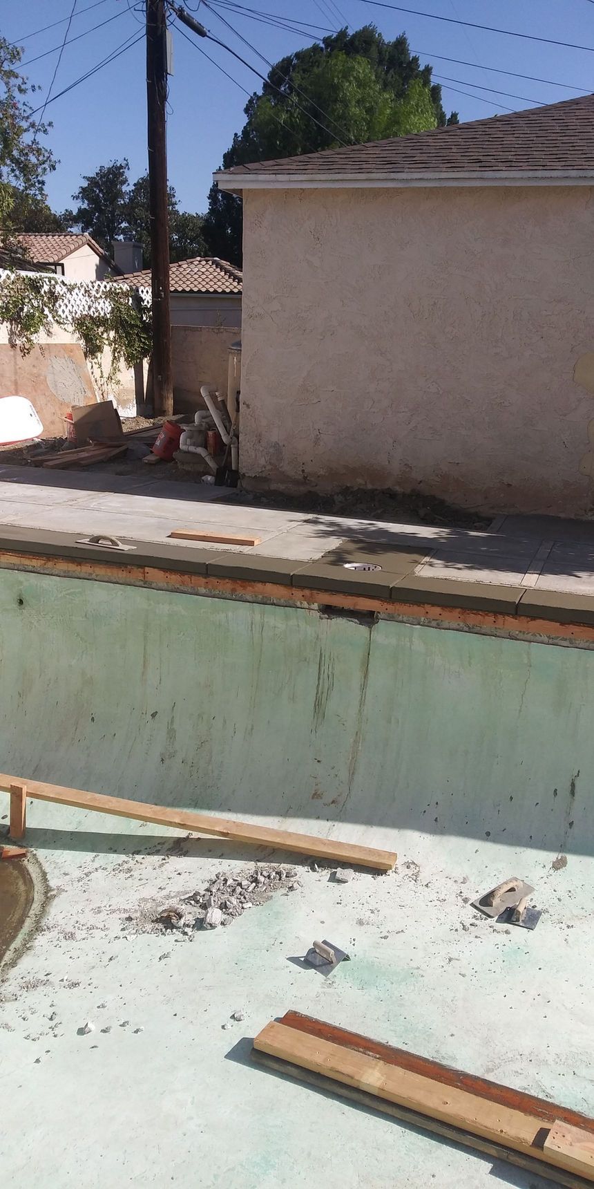 Empty empty pool beside a beige house, with wooden planks and scattered debris inside.