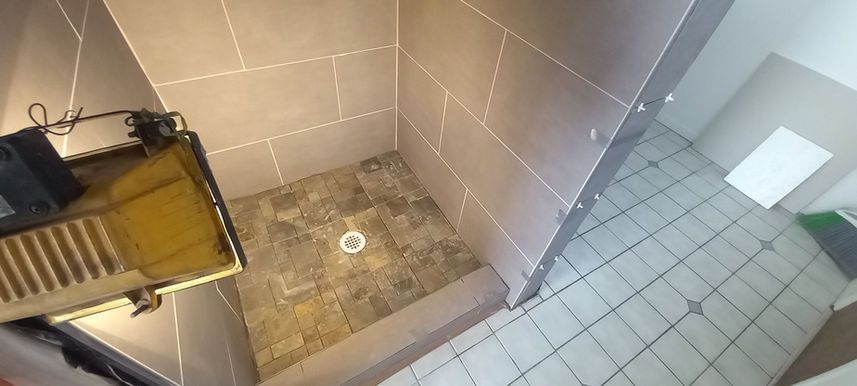 Small tiled shower corner with a drain, beige wall tiles, and a yellow metal cart or chair on the left.