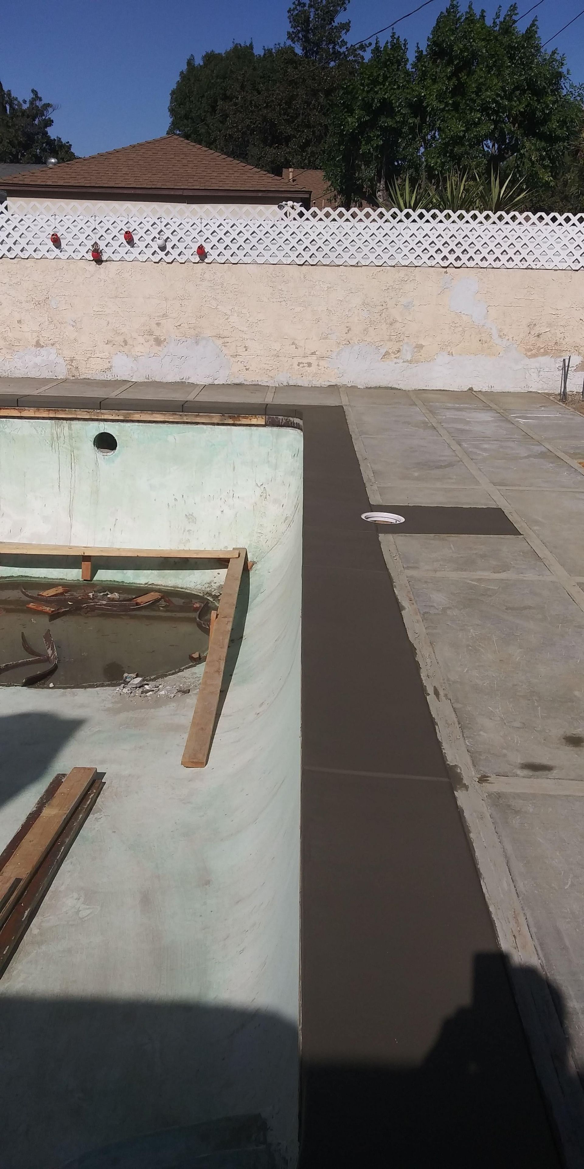 Rooftop with a rectangular pool under construction, concrete deck, and white wall with trees beyond.