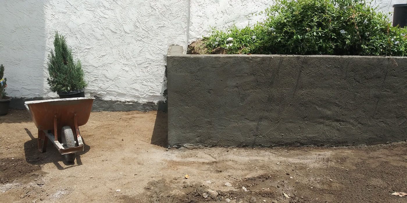 Small wheelbarrow in a sunny courtyard beside a concrete planter wall and white building.