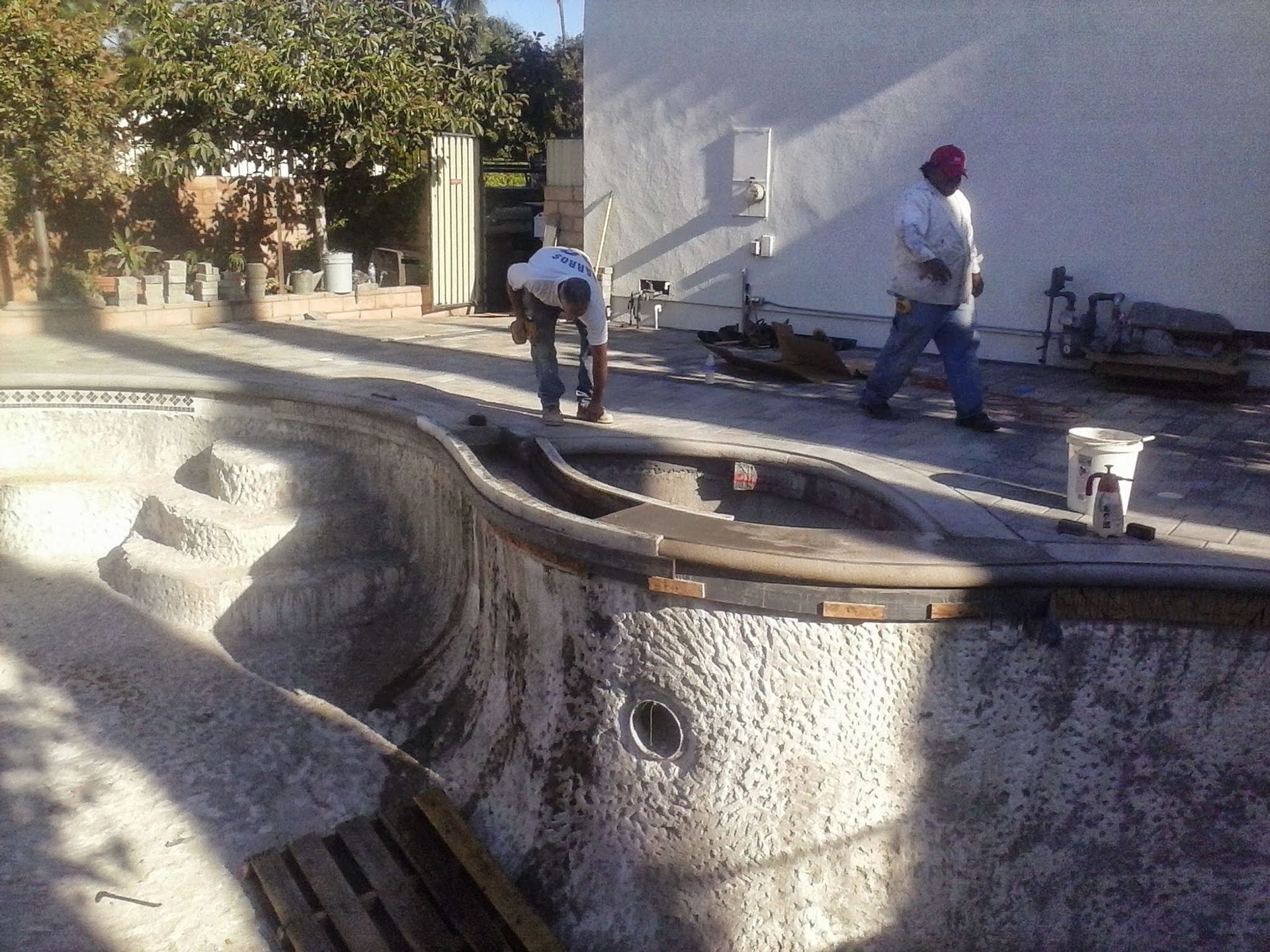 Two workers repair a drained concrete pool beside a building under bright sunlight.
