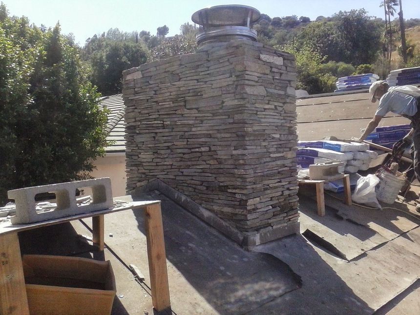 Stone chimney being built on a rooftop with tools and construction materials around it