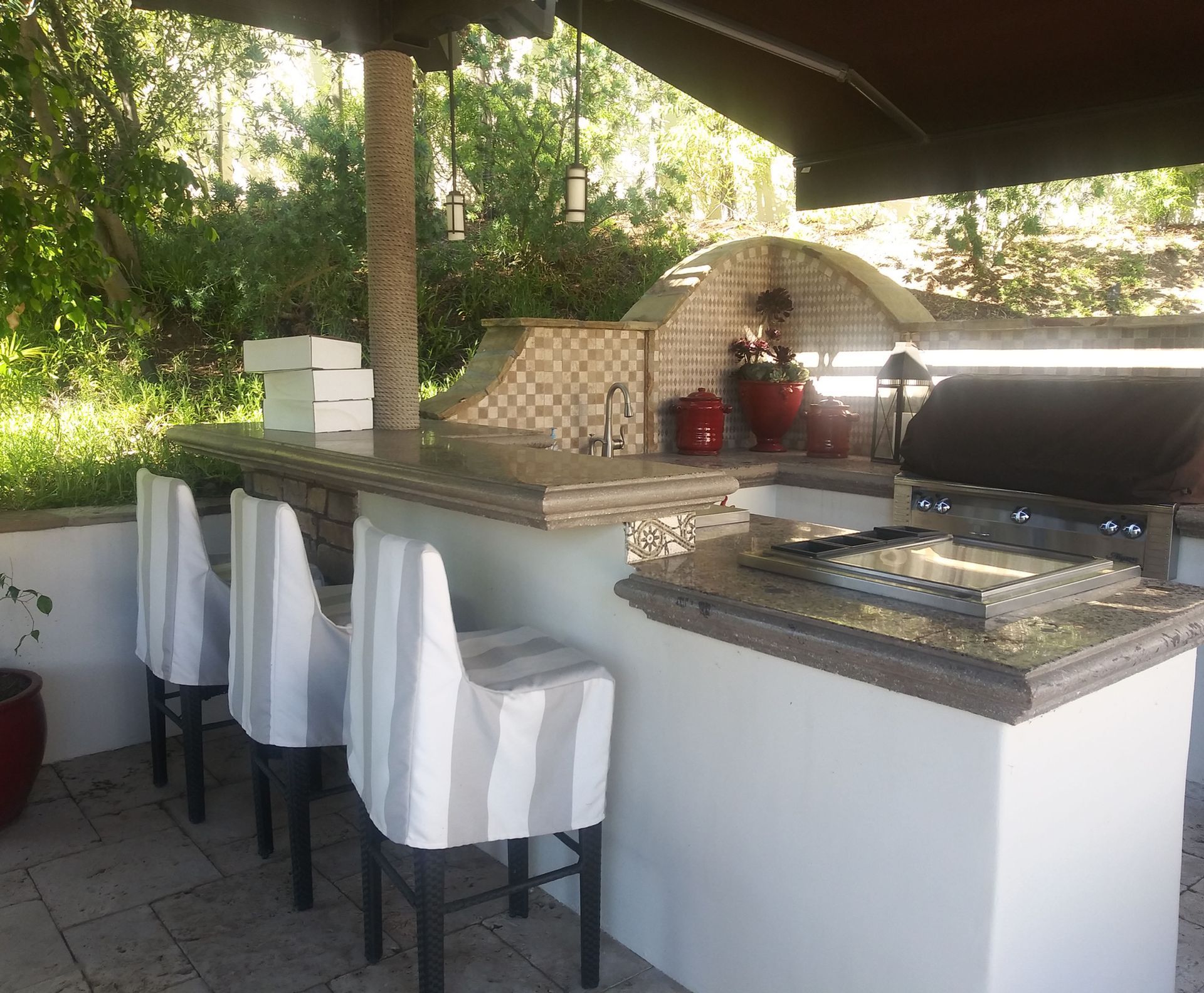Outdoor patio bar with white stools, granite countertop, built-in grill, and pizza oven under a shaded pergola