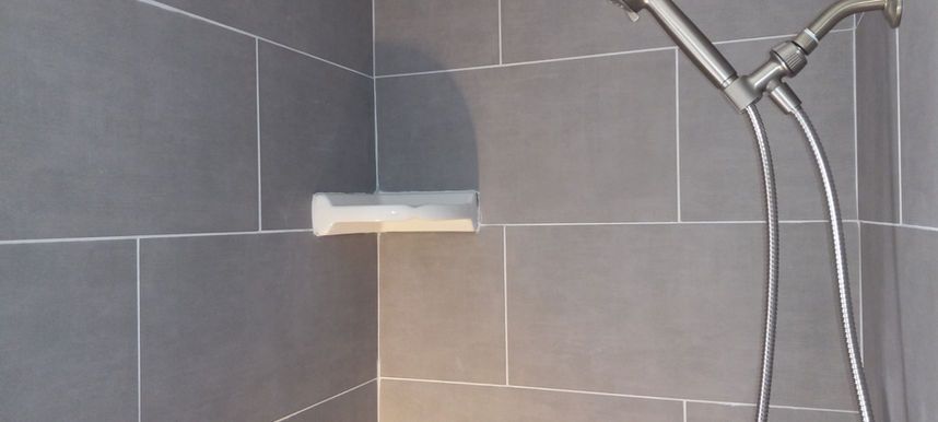 Corner shower with gray tile, a white soap shelf, and chrome showerhead hose