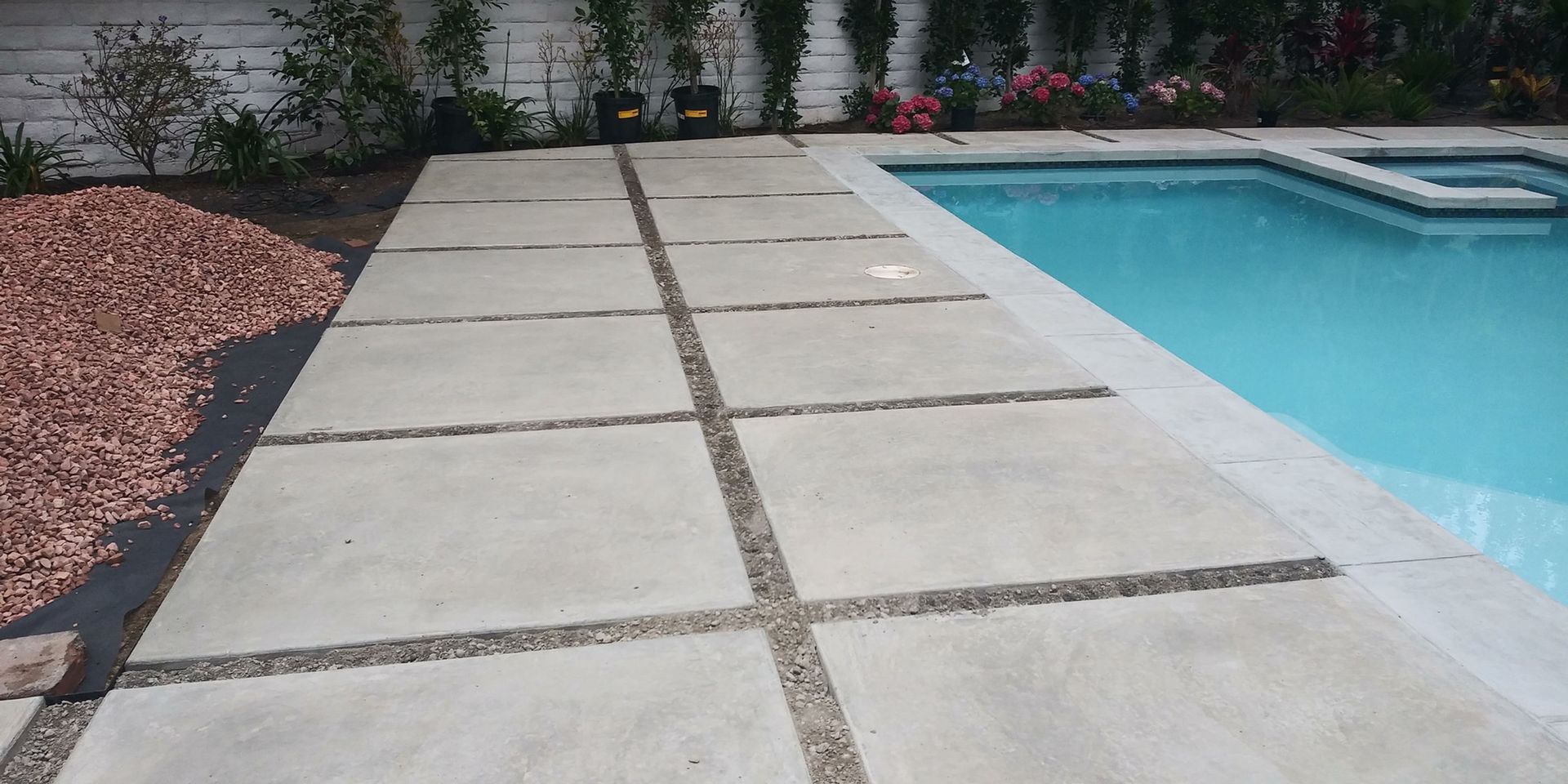 Concrete patio beside a swimming pool with gravel landscaping and plants along the edge