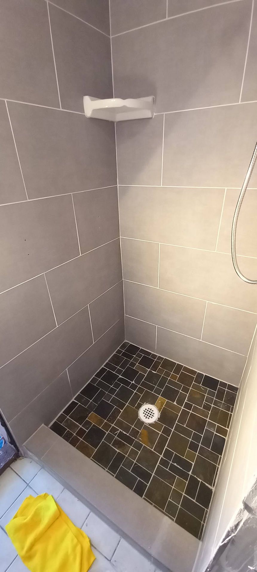 Tiled shower stall with a small shelf, dark mosaic floor, drain, and a yellow towel on the floor.