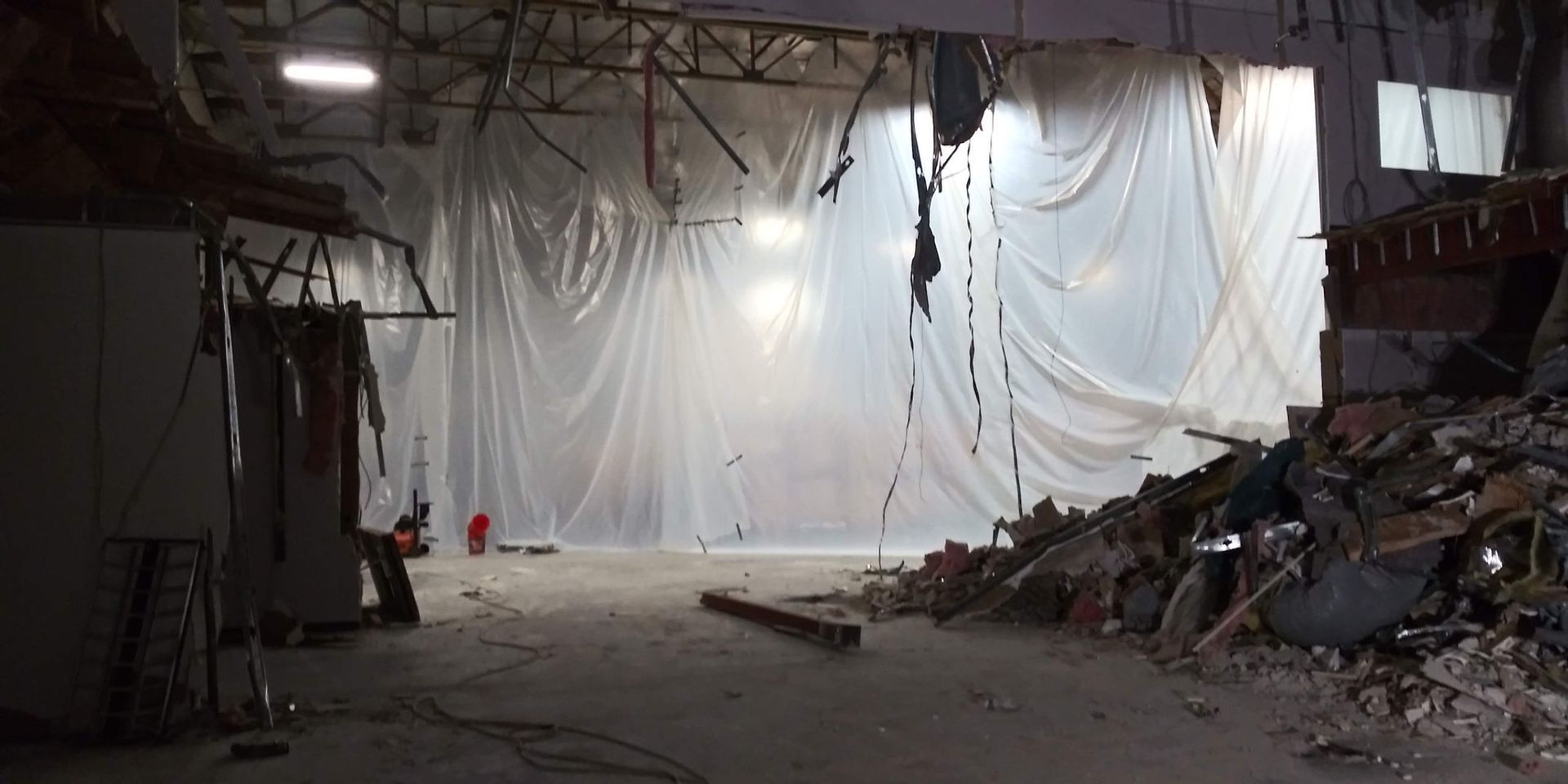 Collapsed industrial interior with rubble, torn plastic sheeting, and debris-strewn floor under dim light
