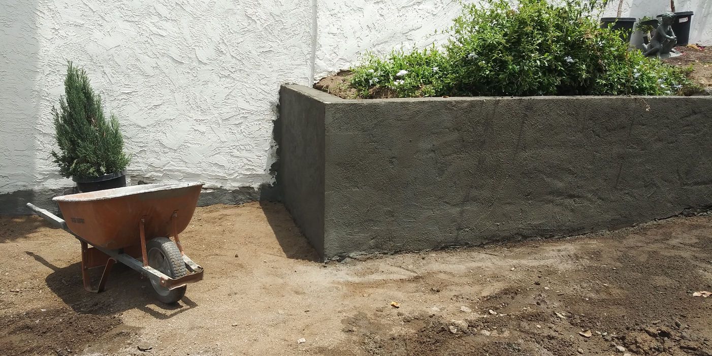 Wheelbarrow in a dirt yard beside a raised planter against a white wall.