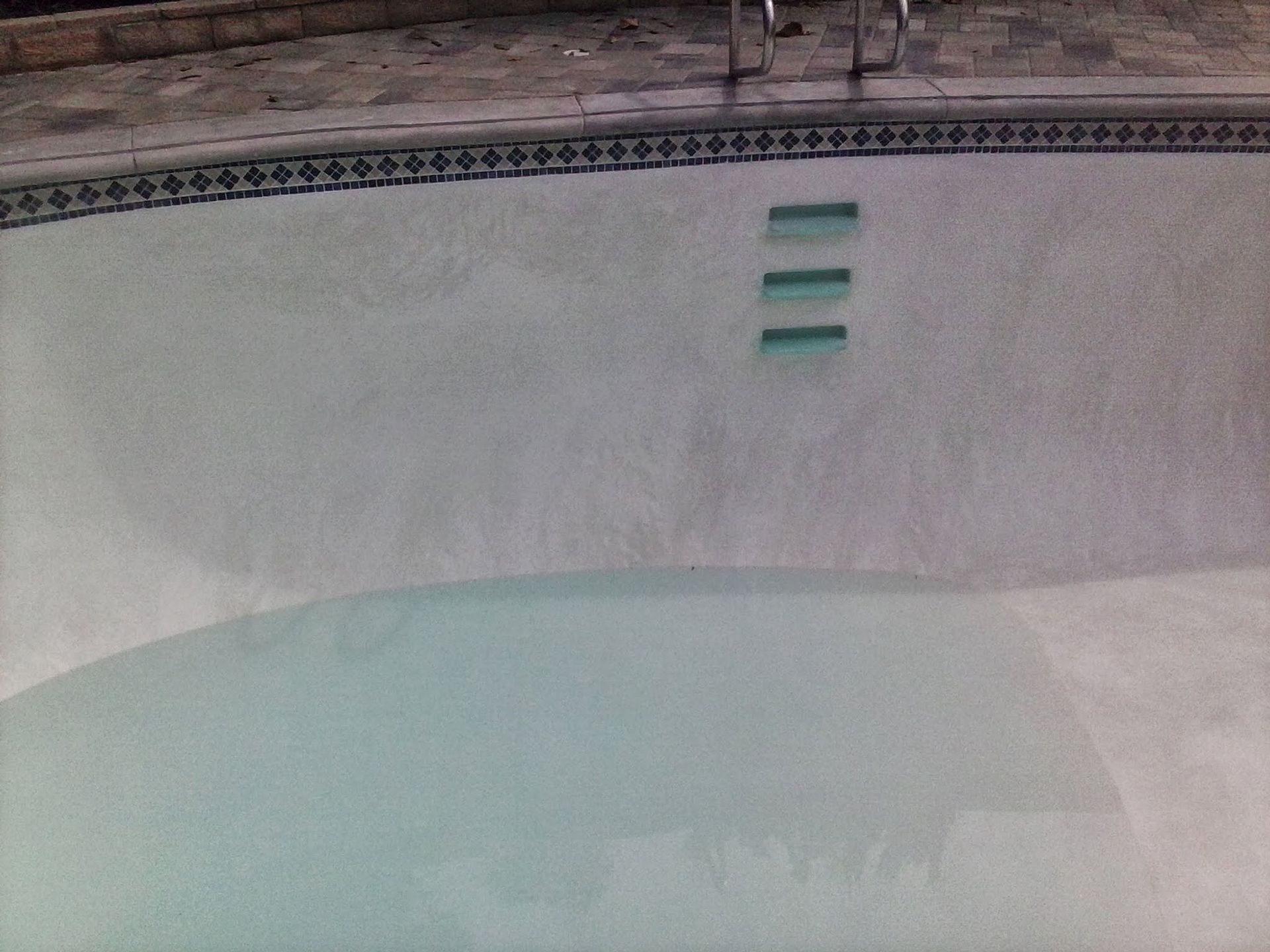 Empty swimming pool with pale blue water and a tiled wall with three green steps/lights.