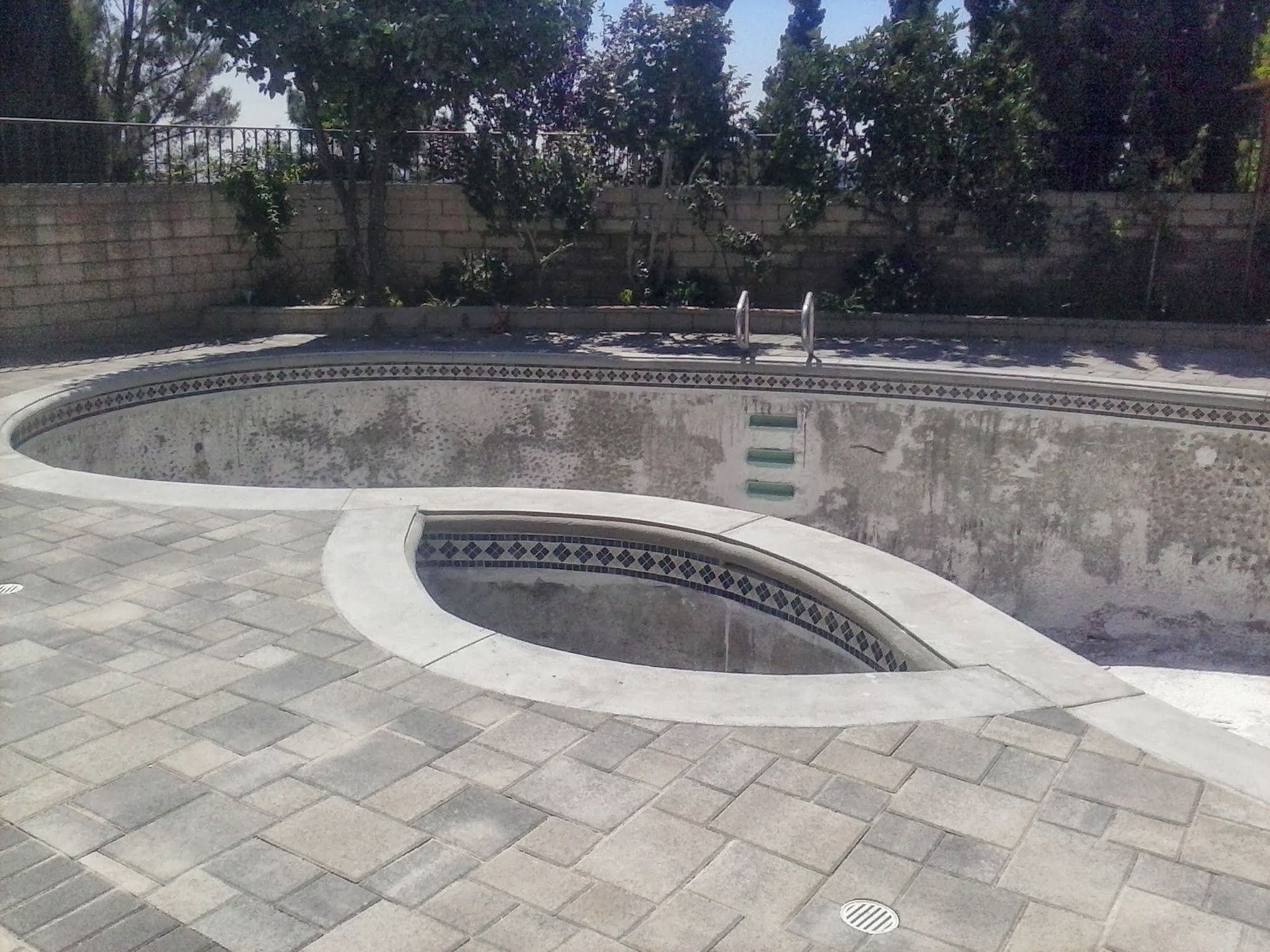 Empty empty swimming pool with a curved spa, tiled deck, and trees in the background