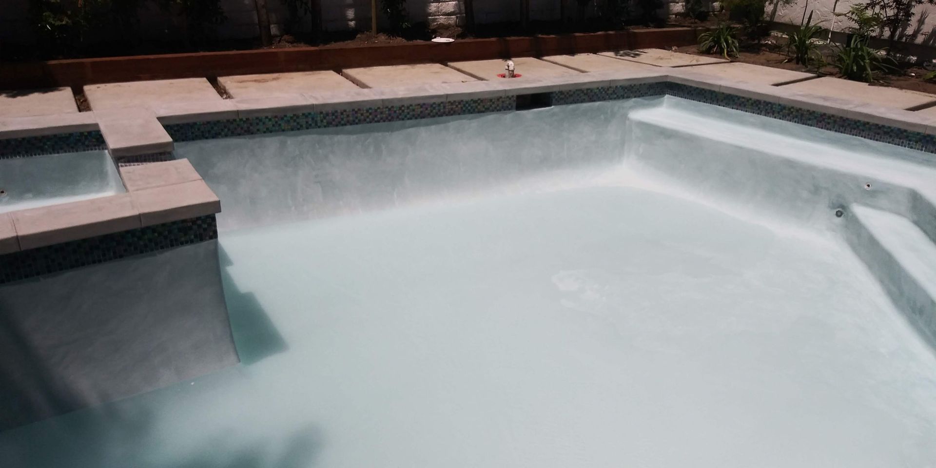 Empty backyard swimming pool with cloudy water and tan tile coping