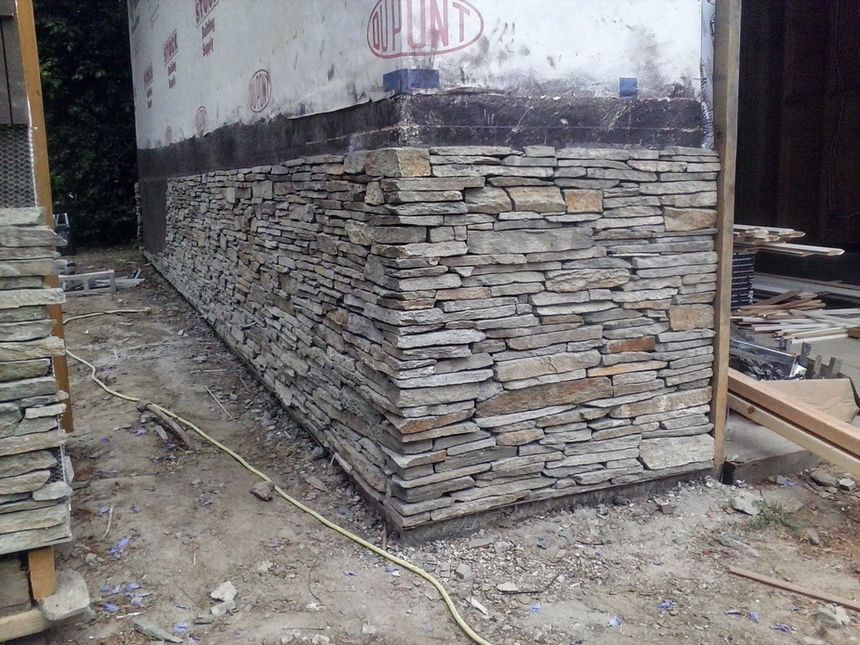 Stone-clad building corner under construction with exposed upper wall and dirt ground around it