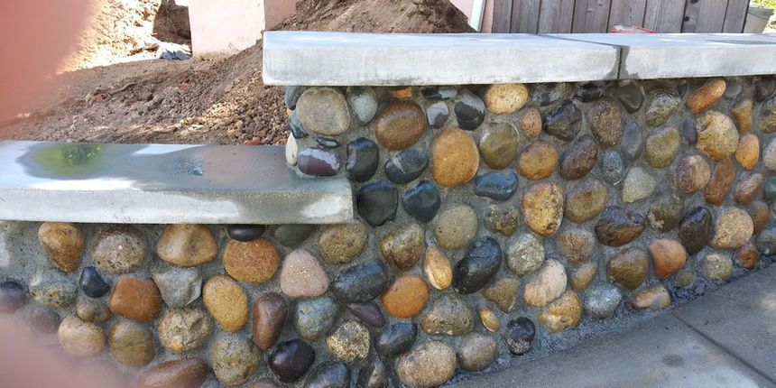 Stone retaining wall with multicolored river rocks and a concrete cap beside a dirt path