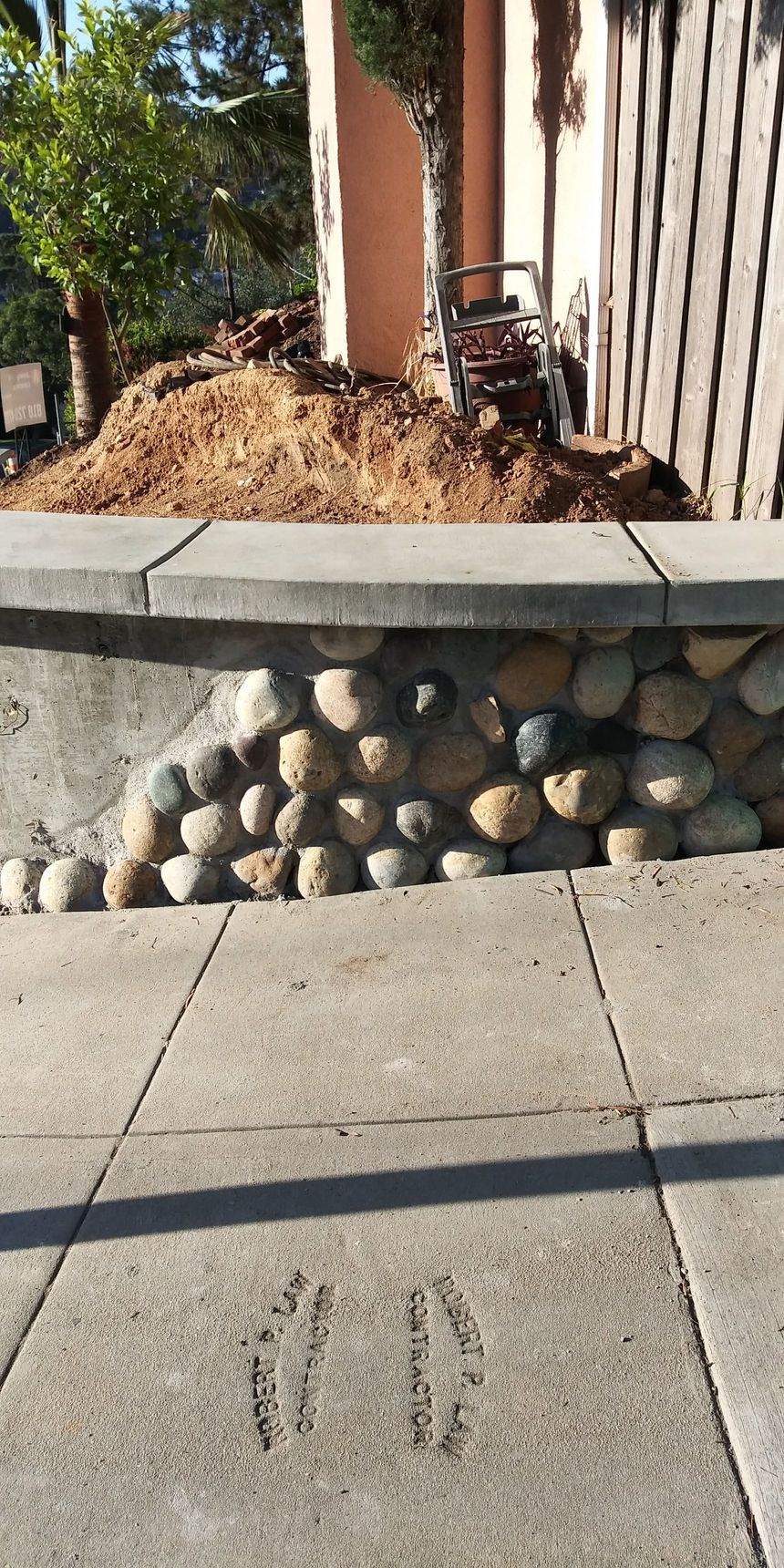 Sidewalk with long shadows in front of a stone retaining wall and a landscaped planter with mulch and a tree