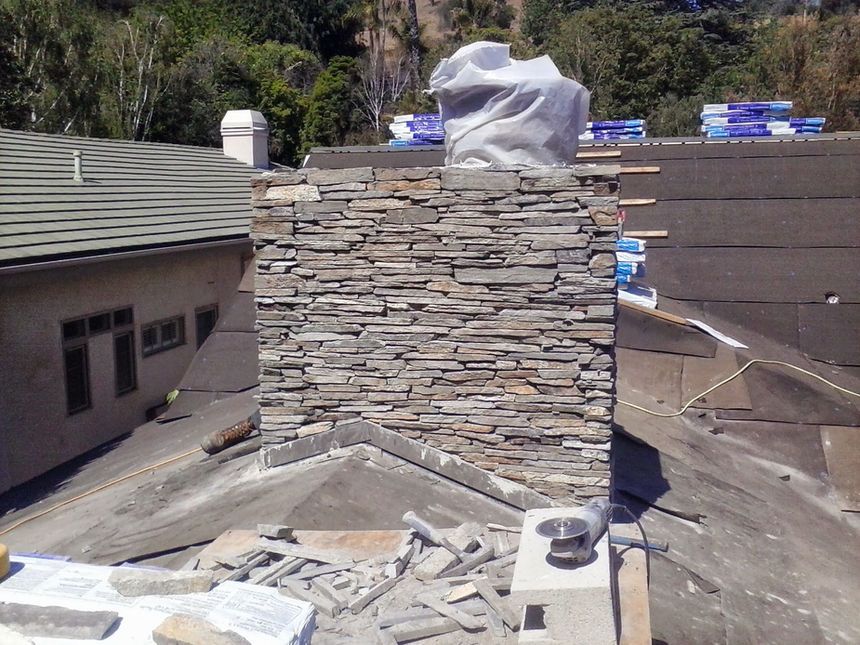 Rooftop with a stone chimney under construction, surrounded by roofing materials and nearby houses.