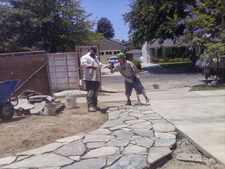 Two people standing on a stone walkway in a sunny residential driveway, one holding a tool and gesturing.