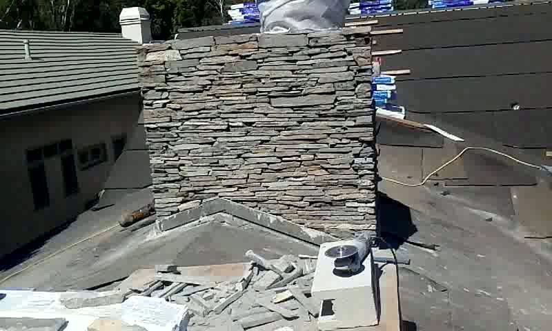 Rooftop chimney clad in stacked stone, with roofing materials and debris scattered around it.