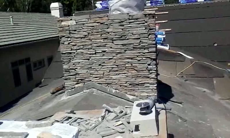 Rooftop stone chimney under construction with gray shingles and scattered tools and debris