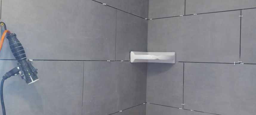 Tiled shower corner with a wall-mounted soap holder and a blue handheld shower hose on the left