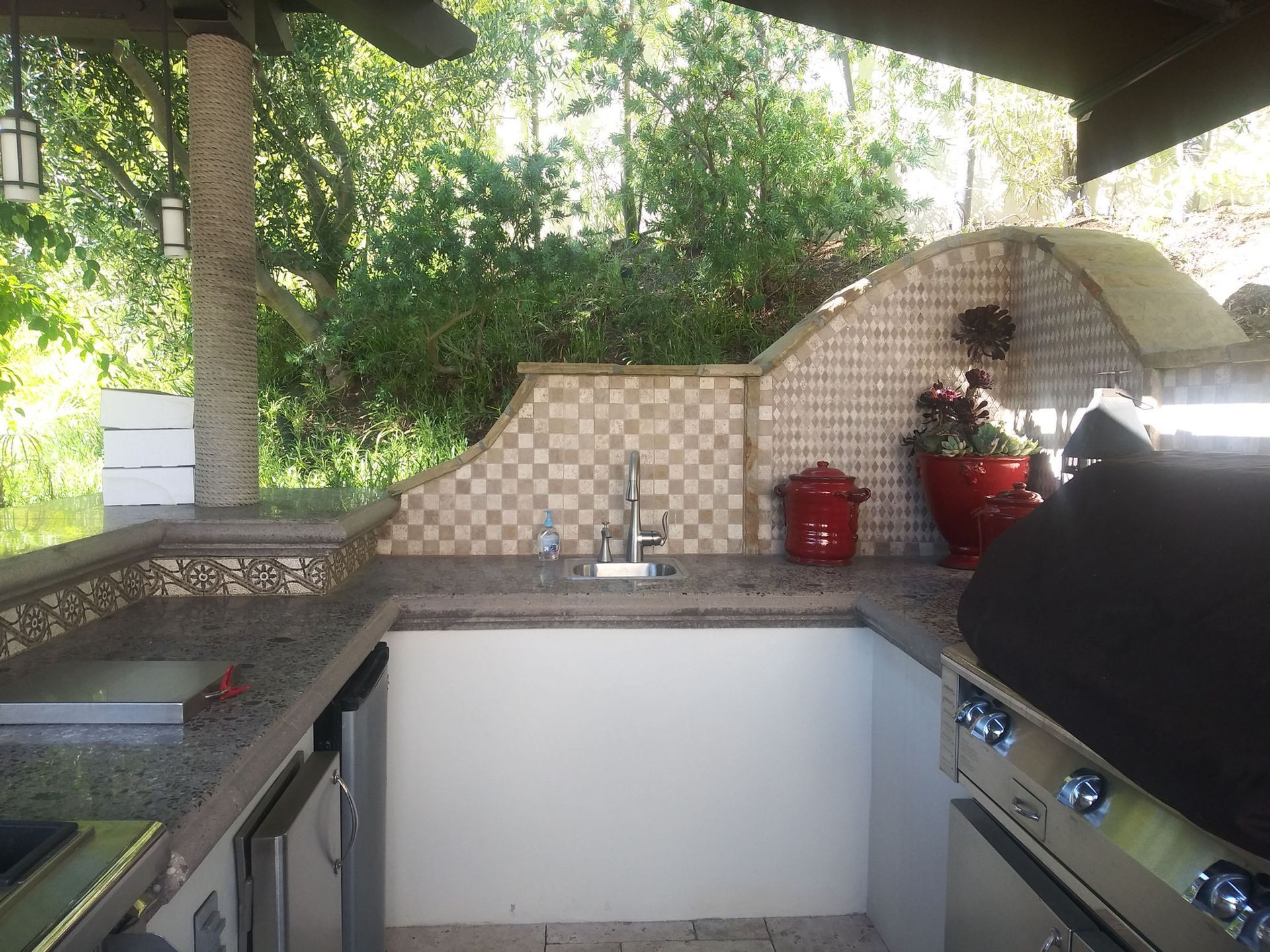 Outdoor kitchen with tiled counter, sink, grill, and red pots under a covered patio