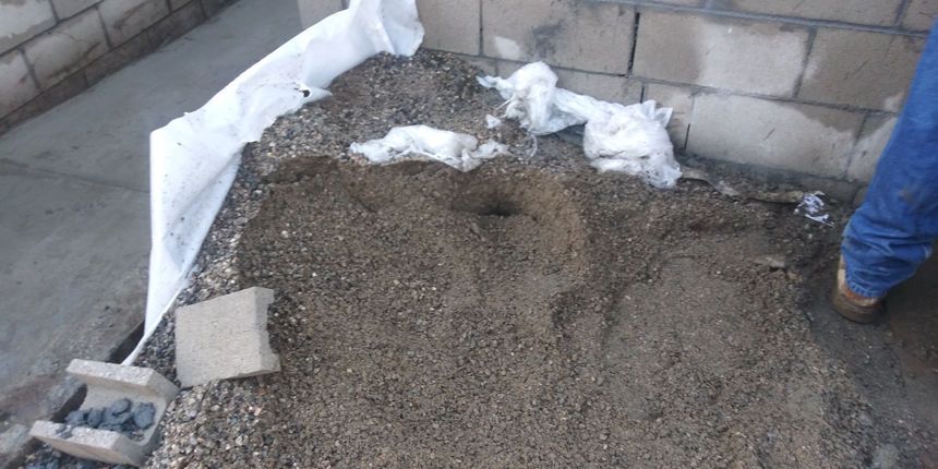 Pile of wet gravel and cement mix beside a cinder block wall, with a workerβs legs visible on the right